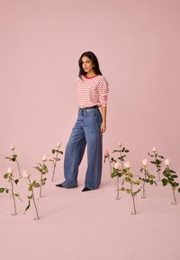 Noor naine, kes kannab punaseid ja valgeid triibulisi särki ning laia siniseid teksapükse, seisab roosade rooside seas, mis on selgetes vaasides roosa tausta ees.