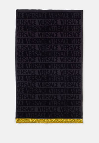Serviette noire avec le texte en relief "VERSACE" répété et une bordure jaune décorative à motifs sur le bord inférieur.