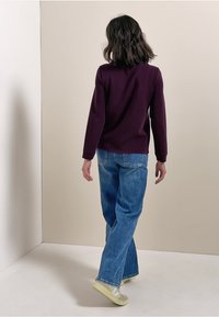 Haut violet à manches longues, coupe décontractée, associé à un jean taille haute bleu clair. Le mannequin porte des baskets de couleur claire, les cheveux coiffés de manière lâche.