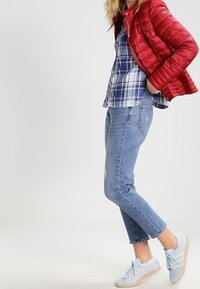 Rød pufferjakke, blå ternet skjorte, lyseblå jeans og blå sneakers. Bemærk den quiltede struktur og den manchetede kant på jeansene.