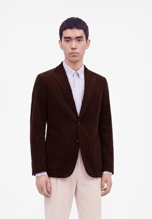 Jeune homme portant un blazer en velours côtelé brun foncé sur une chemise lavande claire à boutons et un pantalon beige clair sur fond blanc.
