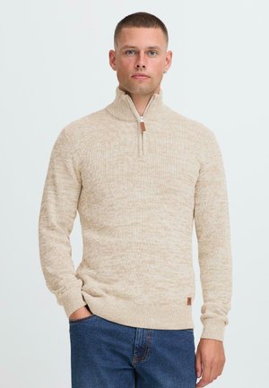 Hombre con cabello corto que lleva un suéter de punto beige con cremallera de un cuarto y jeans azules, de pie contra un fondo claro y liso.