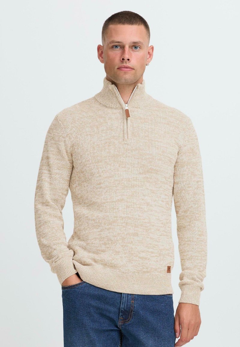 Mann mit kurzem Haar, der einen beigen Strickpullover mit einem Viertel-Reißverschluss und blaue Jeans trägt, steht vor einem schlichten hellen Hintergrund.