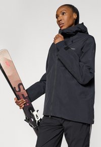 Sort vandtæt skijakke med zip-lukning, høj krave og justerbar hætte, parret med ski med lysebrune og sorte accenter.
