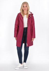 Neslankus bordo spalvos paltas su kapišonu, priekiniais užtrauktukais, šoninėmis kišenėmis ir metalinėmis detalėmis. Derinamas su tamsiomis džinsinėmis kelnėmis ir baltomis sportbatėmis.