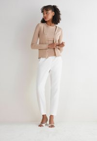 Beżowy kardigan w prążki na dopasowanej bluzce, w zestawieniu z białymi spodniami i srebrnymi sandałami na obcasie. Gładka tekstura, dopasowany fason, zapięcie na guziki.