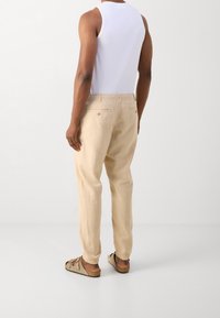 Beige linnen broek met elastische boorden en achterzakken, gedragen met een witte tanktop en bruine sandalen, gezien van achteren.
