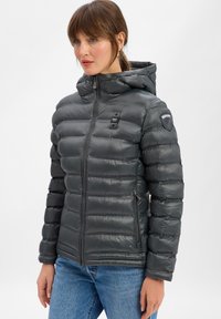 Vrouw in een donkergrijze gewatteerde hooded jas met rits, staand tegen een eenvoudige witte achtergrond, met de handen ontspannen langs haar zij.