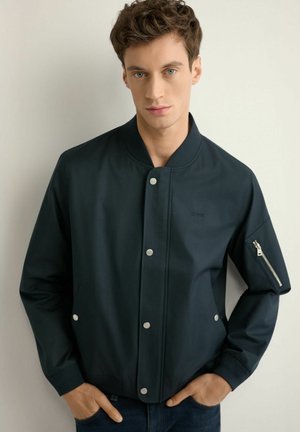 Młody mężczyzna z krótkimi kręconymi włosami, ubrany w granatową kurtkę bomber z srebrnymi zatrzaskami i kieszenią na rękawie na zamek, stojący na tle jasnej ściany.