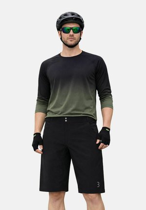 Mann trägt ein schwarzes und grünes Langarm-Radtrikot, schwarze Shorts, fingerlose Handschuhe, Helm und reflektierende Sonnenbrille und steht frontal.