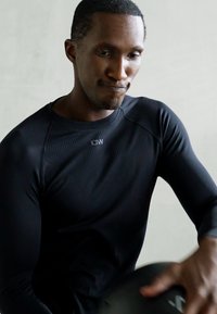 Muškarac u crnoj sportskog majici drži medicinsku loptu, koncentrisan i angažovan u workout aktivnosti na jednostavnoj pozadini.