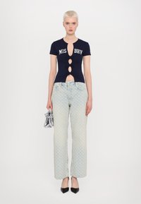 Navy short-sleeve top with cutout details and brand lettering, paired with light blue patterned jeans and a metallic silver handbag.
