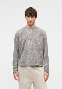 Noor mees, kes kannab halli tumesinise kamuflaažimustriga pikka varrukat särki ja heledaid beežikirjusid pükse, seisab valge taustaga seina ees.