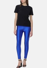 Vrouw in een zwart shirt met korte mouwen, glanzende koningsblauwe leggings en zwarte high heels met enkelbandjes voor een witte achtergrond.