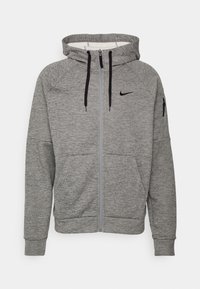 Sweat à capuche gris zippé avec cordons noirs, poches avant, petite poche zippée sur la manche gauche, et logo Nike sur la poitrine.