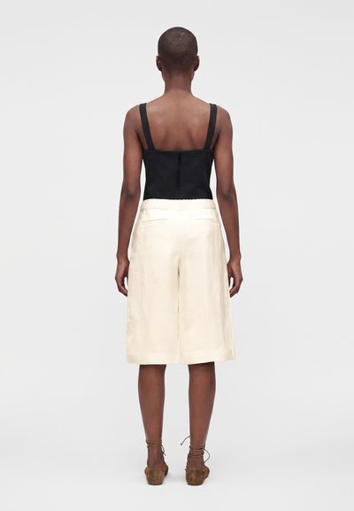 Personne aux cheveux courts portant un haut noir sans manches et un short ample blanc cassé jusqu'aux genoux, debout de dos sur un fond uni.