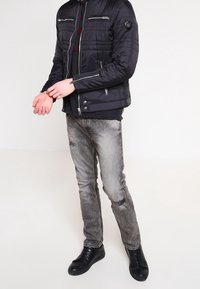 Schwarze gepolsterte Jacke mit Reißverschlussdetails, getragen über einem schwarzen Hemd. Graue, zerrissene Jeans und schwarze Schnürschuhe runden das Outfit ab.