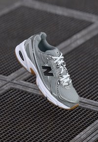 Scarpa da corsa grigia con sovrapposizioni in rete e materiale sintetico, lacci bianchi, dettagli neri e suola in gomma, caratterizzata da un design curvo.