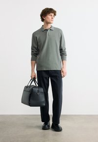 Νέος άνδρας με γκρι μακρυμάνικο πόλο, μαύρο παντελόνι και παπούτσια, κρατώντας μια μαύρη δερμάτινη τσάντα duffel, στέκεται σε γκρι δάπεδο μπροστά σε λευκό τοίχο.