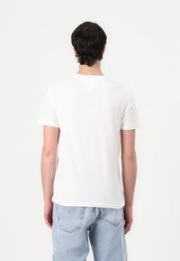 T-shirt en coton blanc à manches courtes et encolure ras du cou, présentant un discret détail de couture en forme de V à l'arrière. Porté avec un jeans en denim bleu clair.