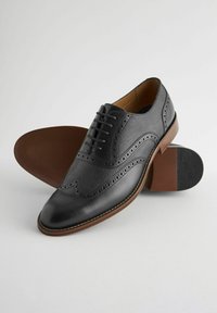 Souliers en cuir noir avec un design brogue, mettant en avant des accents texturés, un laçage à six œillets et une semelle en caoutchouc marron contrastante.