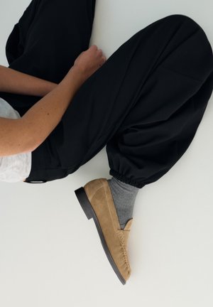 Person, der sidder med bøjede knæ iført sorte løse bukser, grå sokker, lysebrune ruskindspeddelsko og en lysegrå top på en hvid gulv.