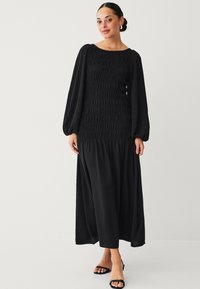 Schwarzes Maxikleid mit langen, puffigen Ärmeln und gerafftem Oberteil. Aus weichem Stoff gefertigt, hat es einen fließenden Rock und einen Quadrat-Ausschnitt.