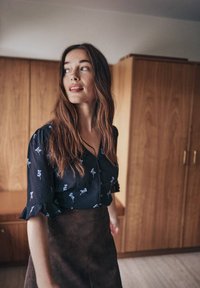 Dunkelbluse mit floralen Motiven und kurzen Rüschensäumen, kombiniert mit einem braunen Wildlederrock. Natürlicher Holz-Hintergrund mit schlichten Möbeln.