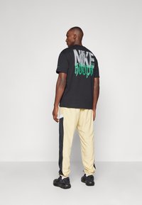Mand iført sort Nike-shirt med grøn "HOOPS" tekst og beige træningsbukser med sorte sidepaneler, står mod en ensfarvet hvid baggrund.