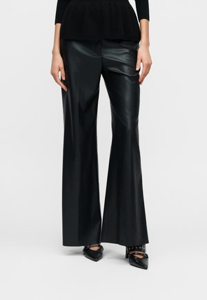 Schwarze weite Hose aus Kunstleder mit glatter Textur, hoher Taille und scharfen Bügelfalten. Kombiniert mit spitzen schwarzen High Heels.