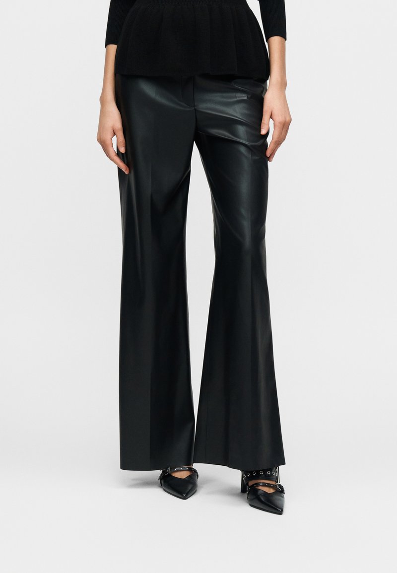 Μαύροι παντελόνες από ψεύτικο δέρμα με φαρδιά γραμμή, λεία υφή, ψηλή μέση και έντονες πιέτες. Συνδυασμένοι με μυτερά μαύρα τακούνια.