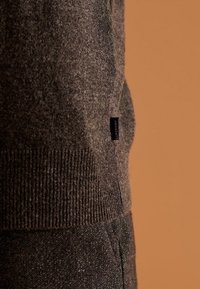 Maglione a maglia grigio scuro con polsini costolati, caratterizzato da un piccolo etichetta blu con la scritta "Superdry." Texture morbida e design aderente.
