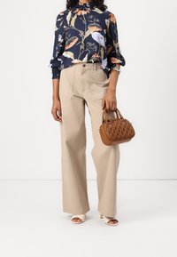 Floral-patterned navy blouse with ruffled neck, beige wide-leg trousers, and a small textured brown handbag. Open-toe heeled shoes.