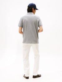 Man in een grijs poloshirt, witte broek, donkergekleurde loafers en een blauwe pet, staand met zijn rug naar de camera tegen een vlakke witte achtergrond.