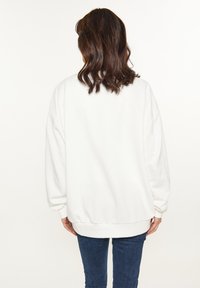 Fehér, oversized pulóver, bordás mandzsettával és alsó szegéllyel. Puha tapintás, laza illeszkedés. Kék farmerral viselve, hátulról nézve.