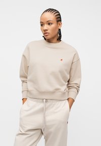 Γυναίκα με πλεξούδες, φορώντας μπεζ φούτερ και αντίστοιχο παντελόνι φόρμας, με τα χέρια στις τσέπες, κοιτάζει προς τα πλάγια μπροστά σε λευκό φόντο.