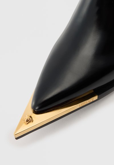 Svart spetsig sko med guldig metallspets som har ett litet Medusa-huvud och Versace-logotypen på sidan.