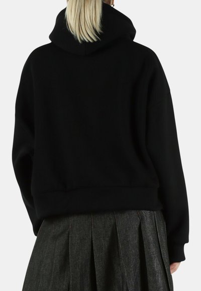 Persoon met blond haar draagt een zwarte capuchontrui en een donker geplooide rok, gezien van achteren tegen een effen achtergrond.