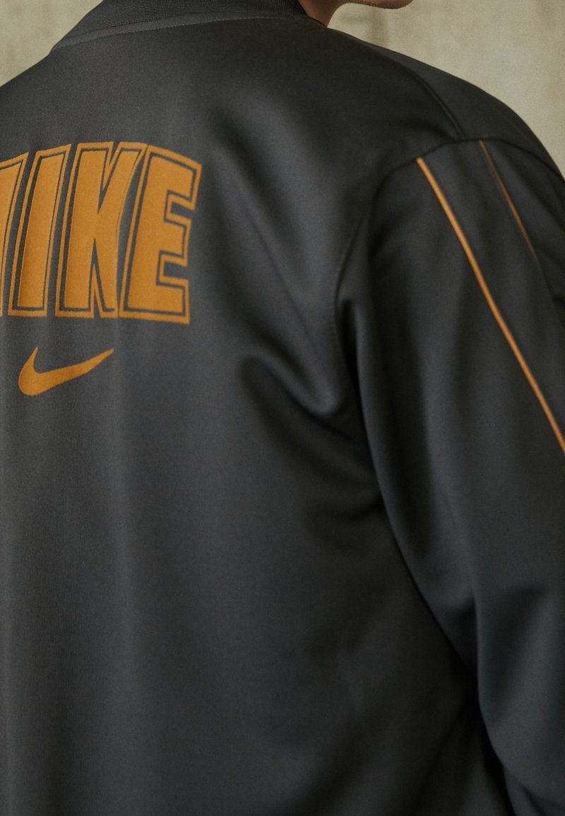 Svart jacka med en slät yta, med djärva orange "NIKE"-bokstäver och logotyp. Designen inkluderar kontrasterande sömmar längs sömmarna.