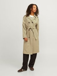 Khakifarbener Trenchcoat mit doppelt geknöpftem Verschluss, Reverskragen und gebundenem Taillengürtel. Kombiniert mit schwarzen Hosen und braunen Sneakers.