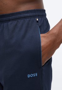 Άνδρας με σκουρόχρωμο δέρμα που φοράει σκούρο μπλε σορτς με ελαστική ζώνη μέσης, με το χέρι στην τσέπη, και το «BOSS» κεντημένο με ανοιχτό μπλε χρώμα στο πόδι.