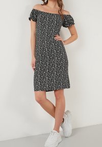 Sort off-the-shoulder kjole med hvidt blomstermønster, korte puffærmer, tætsiddende design, knælang, parret med hvide sneakers.