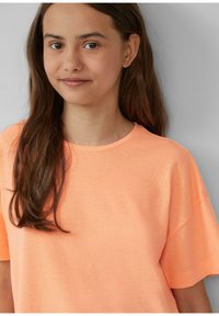 Jong meisje met lang bruin haar, gekleed in een effen perzikkleurig T-shirt met korte mouwen, tegen een lichtgrijze achtergrond.