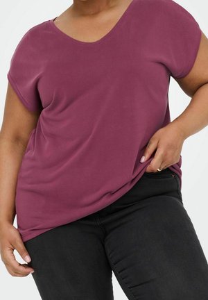 Person trägt ein kurzärmeliges, locker sitzendes lila Oberteil und schwarze Hose, wobei eine Hand am Saum des Shirts zieht.