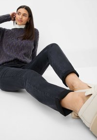 Donkerdenim jeans met opgerolde cuffs, gecombineerd met lichtgekleurde sneakers. De gebreide trui heeft een geometrisch patroon in paars en grijs.