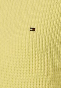 Gros plan sur un tissu tricoté jaune pâle présentant un petit logo brodé rectangulaire en noir, blanc et rouge.