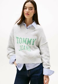 Grauer Strickpullover mit grüner "TOMMY JEANS" Schreibweise, V-Ausschnitt und geripptem Saum, darüber getragen über einem blau-weiß gestreiften Hemd.