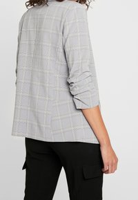 Blazer à carreaux gris avec une texture douce, doté d'une coupe décontractée, de manches retroussées et d'une fente au dos pour une meilleure liberté de mouvement.