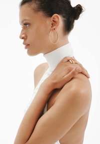 Una modelo lleva una blusa blanca de cuello alto y acanalada. Pendientes de aro dorados y un anillo dorado realzan el conjunto. La piel es suave, mostrando tonos cálidos.