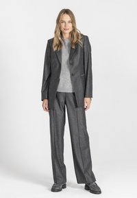 Szary garnitur skrojony na miarę, z marynarką i szerokimi spodniami. Materiał ma teksturowane wykończenie, które uzupełnia jasnoszary sweter noszony pod spodem.